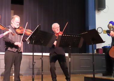 four musicians, three standing and one sitting, playing their instruments