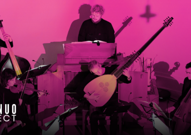 A group of musicians playing period instruments, including a lute, viola da gamba, and a double bass, with an organist in the background. The scene is lit with vibrant pink lighting.