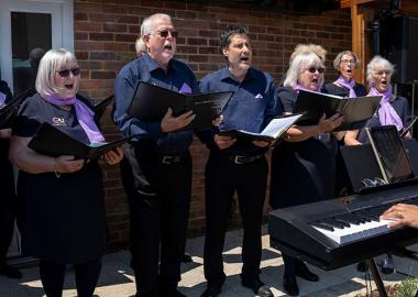 Photo of man playing keyboard and members of OutSingCancer choir singing outdoors on a sunny day.