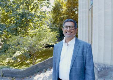 Samir Savant, CEO of St George's Bristol, standing outside on top of some stairs with some pillars and greenery in the background. 