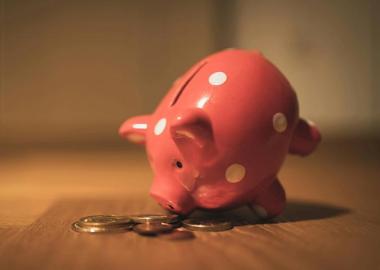 Close-up of a pink piggy bank with white polka dots on a wooden surface. A few coins are scattered in front of it.