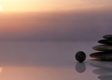 A serene sunset over calm water with a stack of smooth balancing stones reflecting in the water, evoking peace and tranquillity.